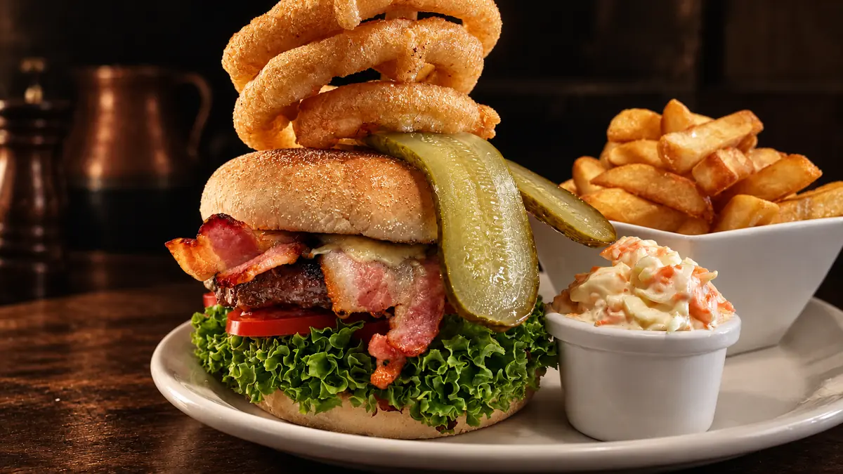 A bacon cheeseburger topped with onion rings and a gherkin, with chips and coleslaw